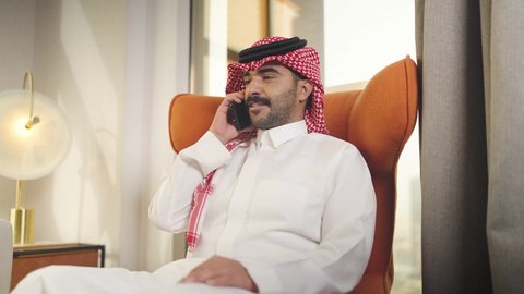 Making a phone call on a mobile phone, chatting online, integrating advanced technologies into daily life, browsing social media, a young Saudi Arabian Gulf man wearing traditional clothing and a keffiyeh holding a mobile phone, sitting on a comfortable chair in a hotel room, professions and businesses in the Kingdom of Saudi Arabia.