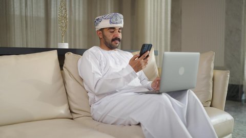 ادارة الأعمال عن بعد، اجراء مكالمه هاتفيه، دمج التقنيات المتطوره فى الحياه اليوميه ، إنجاز المهام المطلوبة عبر اللابتوب، رجل عربي خليجي سعودي يرتدي الزى التقليدي مع الشماغ يجلس على اريكه مريحه في غرفة فندقيه ويستخدم جهاز الحاسوب المحمول والجوال ،التطور والتكنولوجيا الحديثة،  وظائف ومهن سعودية