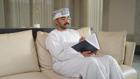 تنمية المهارات الفكرية ، الاهتمام بالعلم والثقافة والدين ، قضاء الوقت بالمطالعة ،ممارسة الهوايات المفضلة،  تعزيز مفهوم المعرفة ،  رجل عربي خليجي عماني يرتدي الدشداشه والكمه يجلس على كرسي مريح فى غرفة فندقيه ويقرأ كتاب ،  الاستمتاع بالهدوء والراحة داخل المنزل