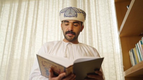 Interest in science, culture, and religion, practicing favorite hobbies, developing intellectual skills, spending time reading, and enhancing the concept of knowledge. An Arab Gulf Omani man wearing a dishdasha and a cloak stands next to his wooden desk, reading a book, enjoying the peace and comfort of home.