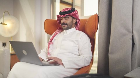 دمج التقنيات المتطوره فى الحياه اليوميه ،  إنجاز المهام المطلوبة عبر اللابتوب، التطور والتكنولوجيا الحديثة، ادارة الأعمال عن بعد،  رجل عربي خليجي سعودي يرتدي الزى التقليدي مع الشماغ يجلس على كرسي مريح في غرفة فندقيه ويستخدم جهاز الحاسوب المحمول ، وظائف ومهن سعودية