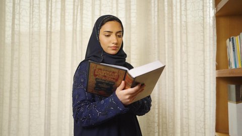 Enhancing the concept of knowledge, practicing favorite hobbies, interest in science, culture, and religion, spending time reading, developing intellectual skills. An Omani Gulf Arab woman wearing an abaya and hijab stands next to a wooden desk, reading a book, enjoying the peace and comfort of home.