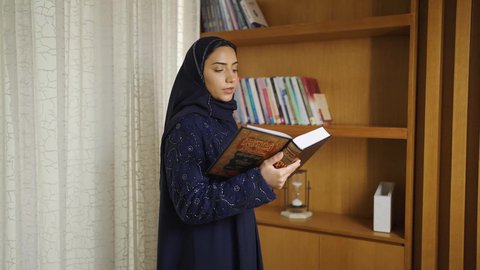 Interest in science, culture, and religion, developing intellectual skills, spending time reading, practicing favorite hobbies, and enhancing the concept of knowledge. An Omani Gulf Arab woman wearing an abaya and hijab stands next to a wooden desk, reading a book, enjoying the peace and comfort of home.