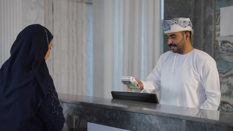 إستعلامات وإستفسارات فى خدمة العملاء ، إجراءات الحجز والدخول لدى مكتب الاستقبال في الفندق، سياحه وفندقه وسفر، رجل عربي خليجي عماني  يعمل في مجال الإستعلامات ، سيده عربية خليجية عمانيه  ترتدي العباءه والحجاب تقوم بالدفع عند الكاشير بطريقة الكترونية بإستخدام بطاقة الإئتمان البنكية