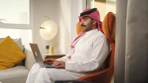 إنجاز المهام المطلوبة عبر اللابتوب، دمج التقنيات المتطوره فى الحياه اليوميه ، التطور والتكنولوجيا الحديثة، ادارة الأعمال عن بعد،  رجل عربي خليجي سعودي يرتدي الزى التقليدي مع الشماغ يجلس على كرسي مريح في غرفة فندقيه ويستخدم جهاز الحاسوب المحمول ، وظائف ومهن سعودية