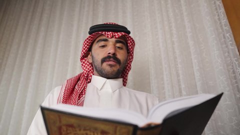 تنمية المهارات الفكرية ، الاهتمام بالعلم والثقافة والدين ، تعزيز مفهوم المعرفة ، قضاء الوقت بالمطالعة ، ممارسة الهوايات المفضلة،رجل عربي خليجي سعودي يرتدي الزى التقليدي مع الشماغ يقف بجانب مكتبه خشبيه ، يأخذ منها كتاب ويبدأ بالقراءه ،  الاستمتاع بالهدوء والراحة داخل المنزل