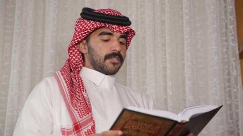 Promoting the concept of knowledge, developing intellectual skills, interest in science, culture, and religion, spending time reading, practicing favorite hobbies. A Saudi Arabian Gulf man wearing traditional clothing with a shemagh stands next to his wooden desk, takes a book from it, and begins reading. Enjoying the peace and comfort of home.