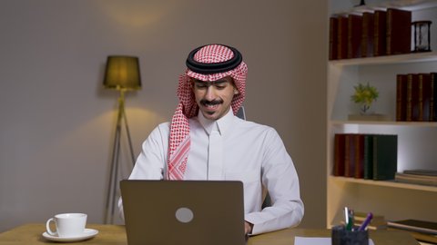 إنجاز المهام المطلوبة عبر اللابتوب، تصوير مقرب لرجل عربي خليجي سعودي يرتدي الزى التقليدي مع الشماغ يجلس على كرسي في غرفة المعيشه ويستخدم جهاز الحاسوب المحمول ،التطور والتكنولوجيا الحديثة، ادارة الأعمال عن بعد، استخدام جهاز اللابتوب ،دمج التقنيات المتطوره فى الحياه اليوميه