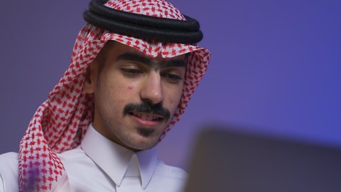 Using a laptop, integrating advanced technologies into daily life, completing tasks via laptop, remote business management, development and modern technology, close-up of a Saudi Arabian Gulf man wearing traditional clothing and a shemagh sitting on a chair in the living room using a laptop.