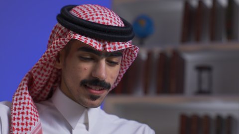 Close-up of a Saudi Arabian Gulf man wearing traditional clothing with a shemagh sitting on a chair in the living room using a laptop, remote business management, development and modern technology, Saudi jobs and professions, integrating advanced technologies into daily life, completing required tasks via laptop, using a laptop