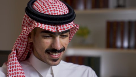 Development and modern technology, Saudi jobs and professions, integrating advanced technologies into daily life, completing required tasks via laptop, remote business management, close-up of a Saudi Arabian Gulf man wearing traditional clothing with a shemagh sitting on a chair in the living room and using a laptop, using a laptop
