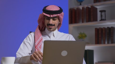 Saudi jobs and professions, drinking hot drinks, integrating advanced technologies into daily life, completing tasks via laptop, remote business management, development and modern technology, a Saudi Arabian Gulf man wearing traditional clothing with a shemagh sitting on a chair in the living room using a laptop, using a laptop.