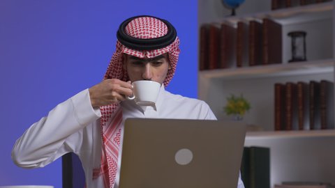 Drinking hot beverages, integrating advanced technologies into daily life, completing tasks via laptop, Saudi jobs and professions, remote business management, development and modern technology, a Saudi Arabian Gulf man wearing traditional clothing with a shemagh sitting on a chair in the living room using a laptop, using a laptop.