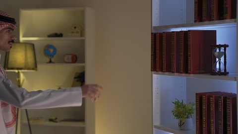 تنمية المهارات الفكرية ،تعزيز مفهوم الثقافة والمعرفة، قضاء الوقت بالمطالعة ، ممارسة الهوايات المفضلة، تصوير من الجانب لرجل عربي خليجي سعودي يرتدي الزى التقليدي مع الشماغ يأخذ كتاب من المكتبه المنزليه ويقوم بقراءة الكتاب ،الاستمتاع بقراءة الروايات والقصص