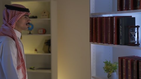 Spending time reading, developing intellectual skills, enhancing the concept of culture and knowledge, practicing favorite hobbies. A side view of a Saudi Arabian Gulf man wearing traditional clothing with a shemagh taking a book from his home library and reading it. Enjoying reading novels and stories.