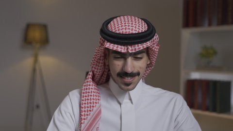 ادارة الأعمال عن بعد،دمج التقنيات المتطوره فى الحياه اليوميه ، التطور والتكنولوجيا الحديثة،  إنجاز المهام المطلوبة عبر اللابتوب، وظائف ومهن سعودية، رجل عربي خليجي سعودي يرتدي الزى التقليدي مع الشماغ يجلس على كرسي في غرفة المعيشه ويستخدم جهاز الحاسوب المحمول ، استخدام جهاز اللابتوب