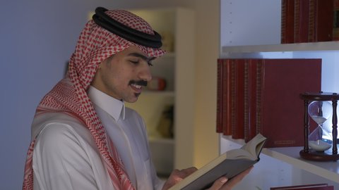 Side view of a Saudi Arabian Gulf man wearing traditional clothing with a shemagh sitting in the living room reading a book. Enjoying reading novels and stories, spending time reading, developing intellectual skills, enjoying peace and comfort inside the home, promoting the concept of culture and knowledge, practicing favorite hobbies.