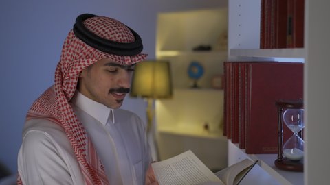 Spending time reading, enjoying the peace and comfort inside the home, enhancing the concept of culture and knowledge, practicing favorite hobbies, developing intellectual skills, enjoying reading novels and stories, a side view of a Saudi Arabian Gulf man wearing traditional clothing with a shemagh sitting in the living room reading a book
