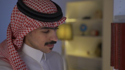 الاستمتاع بالهدوء والراحة داخل المنزل، ممارسة الهوايات المفضلة،  تنمية المهارات الفكرية ، تعزيز مفهوم الثقافة والمعرفة ، قضاء الوقت بالمطالعة ، الاستمتاع بقراءة الروايات والقصص ، تصوير من الجانب لرجل عربي خليجي سعودي يرتدي الزى التقليدي مع الشماغ يجلس في غرفة المعيشة ويقوم بقراءة كتاب