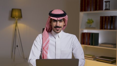 Integrating advanced technologies into daily life, completing required tasks via laptop, Saudi jobs and professions, remote business management, development and modern technology, a Saudi Arabian Gulf man wearing traditional clothing with a shemagh sitting on a chair in the living room using a laptop, using a laptop.