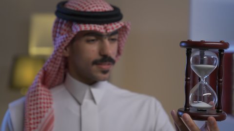 Sand timing control, schedule organization, business management concept, Saudi Arabian Gulf man wearing traditional thobe and shemagh holding glass sand timer (bankam) in his hand looking with happy expression