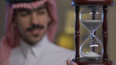 تنظيم الجدول الزمني,  مفهوم إدارة الأعمال ، رجل عربي خليجي سعودي يرتدي الثوب التقليدي والشماغ يمسك بيده الساعة الرملية الزجاجية (البنكام) ينظر بتعابير السعاده, القيام بمراقبة توقيت الرمل