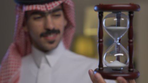 مفهوم إدارة الأعمال وتنظيم الجدول الزمني, رجل عربي خليجي سعودي يرتدي الثوب التقليدي والشماغ يمسك بيده الساعة الرملية الزجاجية (البنكام) ينظر بتعابير السعاده, القيام بمراقبة توقيت الرمل