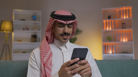 مهن و أعمال في السعودية, الدردشه عبر الانترنت ، اجراء مكالمه فيديو ،التواصل مع العملاء عن بعد، تصفح وسائل التواصل الاجتماعي ، استخدام التقنيات الحديثة و المتطورة في العمل, إجراء اتصال هاتفي عبر الجوال, شاب عربي خليجي سعودي يرتدي الثوب التقليدي و الشماغ  يمسك الهاتف المحمول بيده