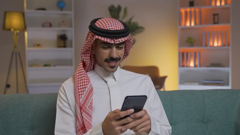 الدردشه عبر الانترنت ، اجراء مكالمه فيديو ،التواصل مع العملاء عن بعد، تصفح وسائل التواصل الاجتماعي ، استخدام التقنيات الحديثة و المتطورة في العمل, إجراء اتصال هاتفي عبر الجوال, مهن و أعمال في السعودية, شاب عربي خليجي سعودي يرتدي الثوب التقليدي و الشماغ  يمسك الهاتف المحمول بيده