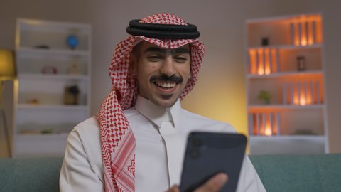 التواصل مع العملاء عن بعد، تصفح وسائل التواصل الاجتماعي ، استخدام التقنيات الحديثة و المتطورة في العمل, إجراء اتصال هاتفي عبر الجوال, مهن و أعمال في السعودية, شاب عربي خليجي سعودي يرتدي الثوب التقليدي و الشماغ  يمسك الهاتف المحمول بيده