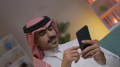 تصفح وسائل التواصل الاجتماعي ، استخدام التقنيات الحديثة و المتطورة في العمل, إجراء اتصال هاتفي عبر الجوال, مهن و أعمال في السعودية, شاب عربي خليجي سعودي يرتدي الثوب التقليدي و الشماغ  يمسك الهاتف المحمول بيده, التواصل مع العملاء عن بعد