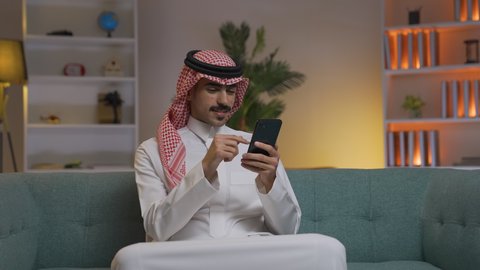Making a phone call via mobile phone, using modern and advanced technologies at work, professions and businesses in Saudi Arabia, a young Saudi Arabian Gulf man wearing traditional thobe and shemagh holding a mobile phone in his hand, communicating with clients remotely, browsing social media