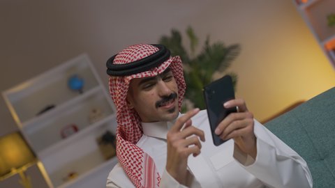 شاب عربي خليجي سعودي يرتدي الثوب التقليدي و الشماغ يمسك الهاتف المحمول بيده، التواصل مع العملاء عن بعد،  تصفح وسائل التواصل الاجتماعي ،إجراء اتصال هاتفي عبر الجوال, مهن و أعمال في السعودية, استخدام التقنيات الحديثة و المتطورة في العمل, الدردشه عبر الانترنت