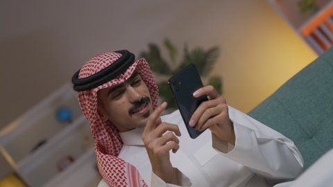 التواصل مع العملاء عن بعد،  تصفح وسائل التواصل الاجتماعي ،إجراء اتصال هاتفي عبر الجوال, مهن و أعمال في السعودية, استخدام التقنيات الحديثة و المتطورة في العمل, الدردشه عبر الانترنت، شاب عربي خليجي سعودي يرتدي الثوب التقليدي و الشماغ يمسك الهاتف المحمول بيده