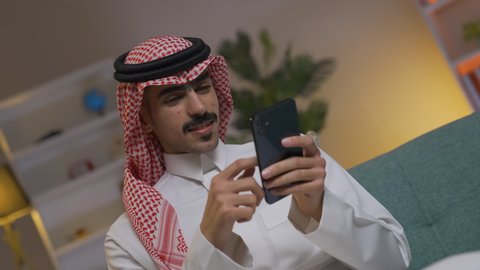 تصفح وسائل التواصل الاجتماعي ،إجراء اتصال هاتفي عبر الجوال, مهن و أعمال في السعودية, التواصل مع العملاء عن بعد، استخدام التقنيات الحديثة و المتطورة في العمل, الدردشه عبر الانترنت، شاب عربي خليجي سعودي يرتدي الثوب التقليدي و الشماغ يمسك الهاتف المحمول بيده