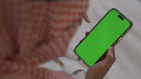 Empty green screen chroma key, young Saudi Arabian Gulf man wearing traditional thobe and shemagh holding mobile phone in hand, browsing social media, making a mobile phone call, jobs and professions in Saudi Arabia, using modern and advanced technologies at work, chatting online