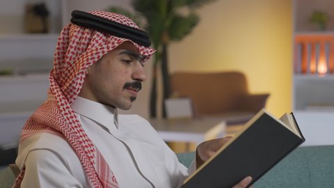 Promoting the concept of culture and knowledge, practicing favorite hobbies, enjoying peace and comfort at home, developing intellectual skills, spending time reading and culture, a side view of a Saudi Arabian Gulf man wearing traditional clothing with a shemagh sitting on the sofa in the living room reading a book, enjoying reading novels and stories