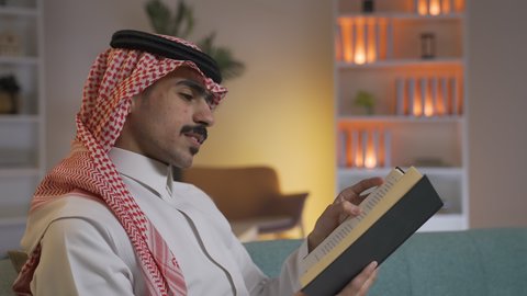 Developing intellectual skills, enjoying peace and comfort at home, spending time reading and learning, practicing favorite hobbies. A side view of a Saudi Arabian Gulf man wearing traditional clothing with a shemagh sitting on a sofa in the living room reading a book. Enjoying reading novels and stories. Reinforcing the concept of culture and knowledge.