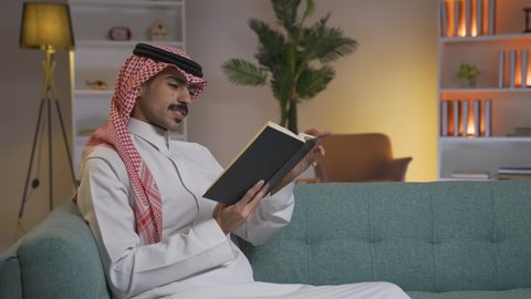 Promoting the concept of culture and knowledge, developing intellectual skills, enjoying peace and comfort at home, spending time reading and cultivating, and practicing favorite hobbies. A Saudi Arabian Gulf man wearing traditional clothing with a shemagh sitting on the sofa in the living room reading a book, enjoying reading novels and stories.