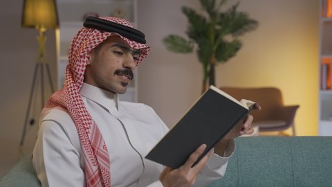 Practicing favorite hobbies, spending time reading and culture, a Saudi Arabian Gulf man wearing traditional clothing with a shemagh sitting on the sofa in the living room reading a book, enjoying reading novels and stories, enhancing the concept of culture and knowledge, developing intellectual skills, enjoying the peace and comfort inside the home