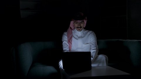 وظائف ومهن سعودية، ادارة الأعمال عن بعد، إنجاز المهام المطلوبة عبر اللابتوب، دمج التقنيات المتطوره فى الحياه اليوميه ، التطور والتكنولوجيا الحديثة، رجل عربي خليجي سعودي يرتدي الزى التقليدي مع الشماغ يجلس على اريكه في غرفة مظلمه ويستخدم جهاز الحاسوب المحمول ، استخدام جهاز اللابتوب