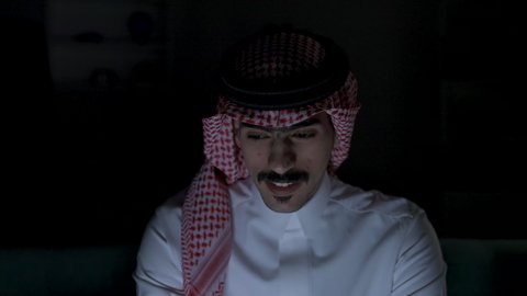 Remote business management, completing required tasks via laptop, integrating advanced technologies into daily life, development and modern technology, Saudi jobs and professions, a Saudi Arabian Gulf man wearing traditional clothing with a keffiyeh sitting on a couch in a dark room using a laptop, using a laptop.