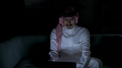Accomplishing required tasks via laptop, integrating advanced technologies into daily life, development and modern technology, Saudi jobs and professions, remote business management, a Saudi Arabian Gulf man wearing traditional clothing with a keffiyeh sitting on a couch in a dark room using a laptop, using a laptop.