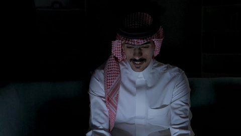 التطور والتكنولوجيا الحديثة،  وظائف ومهن سعودية، ادارة الأعمال عن بعد، إنجاز المهام المطلوبة عبر اللابتوب، رجل عربي خليجي سعودي يرتدي الزى التقليدي مع الشماغ يجلس على اريكه في غرفة مظلمه ويستخدم جهاز الحاسوب المحمول ، دمج التقنيات المتطوره فى الحياه اليوميه ، استخدام جهاز اللابتوب