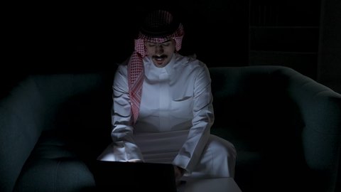 Saudi jobs and professions, remote business management, completing required tasks via laptop, development and modern technology, a Saudi Arabian Gulf man wearing traditional dress with a shemagh sitting on a couch in a dark room using a laptop, integrating advanced technologies into daily life, using a laptop
