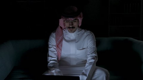 ادارة الأعمال عن بعد، إنجاز المهام المطلوبة عبر اللابتوب، التطور والتكنولوجيا الحديثة، رجل عربي خليجي سعودي يرتدي الزى التقليدي مع الشماغ يجلس على اريكه في غرفة مظلمه ويستخدم جهاز الحاسوب المحمول ، دمج التقنيات المتطوره فى الحياه اليوميه ، استخدام جهاز اللابتوب ، وظائف ومهن سعودية