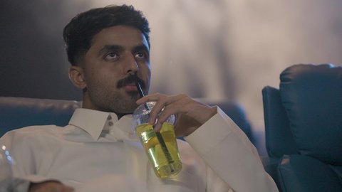 قضاء أوقات مسلية أثناء شرب المشروبات، تصوير مقرب لرجل عربي خليجي سعودي يرتدي الثوب الابيض يجلس داخل السينما يقوم بمشاهدة الفيلم، ايماءات الاندماج و التركيز