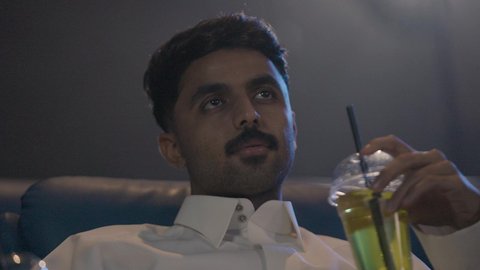 لحظات مرحة مع تناول المشروبات المنعشة، تصوير مقرب لرجل عربي خليجي سعودي يرتدي الثوب الابيض يجلس داخل السينما يقوم بمشاهدة الفيلم، ايماءات الاندماج و التركيز