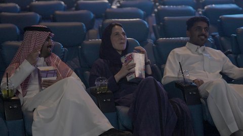 الاستمتاع بتناول المشروبات،  نشاط ترفيهي، دلو الفشار للتسلية ،اصدقاء عربيون خليجيون سعوديون يرتدون الزي التقليدي  يشاهدون فلم في احدى صالات السينما، تعابير وجه و يدين تدل على الحماس و الاستمتاع ، دور العرض
