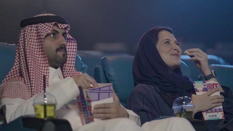 ايماءات الضحك و السرور، دلو الفشار للتسلية ، زوجان عربيان خليجيان سعوديان يرتديان الزي التقليدي يشاهدان فلم في احدى صالات السينما، تعابير وجه و يدين تدل على الحماس و الاستمتاع ، نشاط ترفيهي ، دور العرض، الاستمتاع بتناول المشروبات